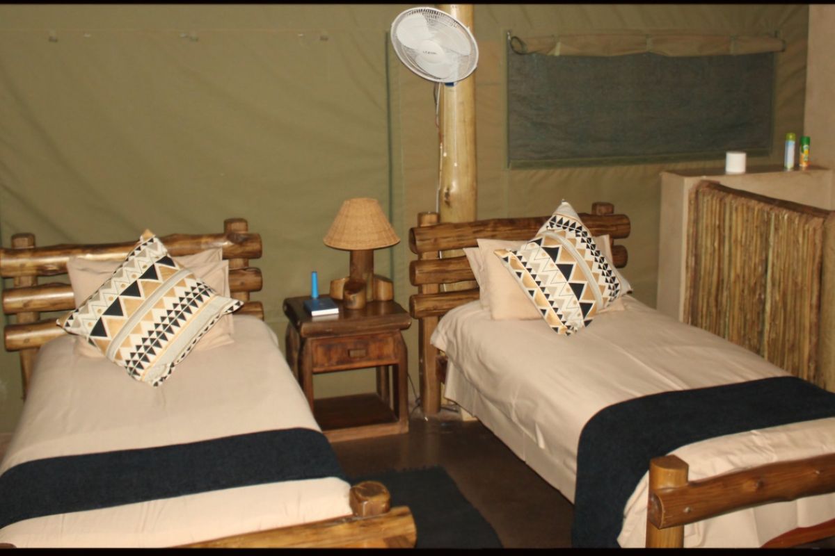 A rustic bedroom featuring canvas walls, twin beds with wooden frames, and a wall-mounted fan within a safari-style tented accommodation.