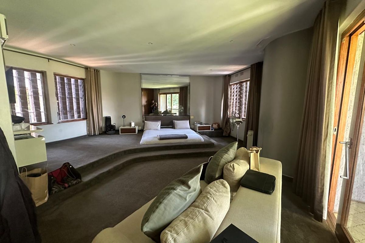 Spacious bedroom featuring a raised platform bed area, large wall mirror, and multiple windows with integrated louvers, offering a unique architectural layout.