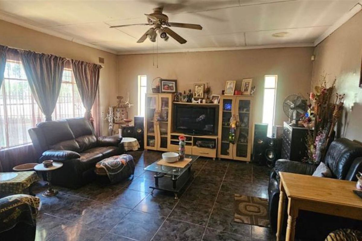 A spacious living room featuring dark floor tiles, a ceiling fan, and large windows with security bars.