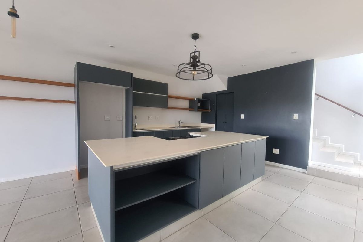 Modern open-plan kitchen featuring a central island, sleek dark cabinetry, stone countertops, and tiled flooring, adjacent to a staircase.
