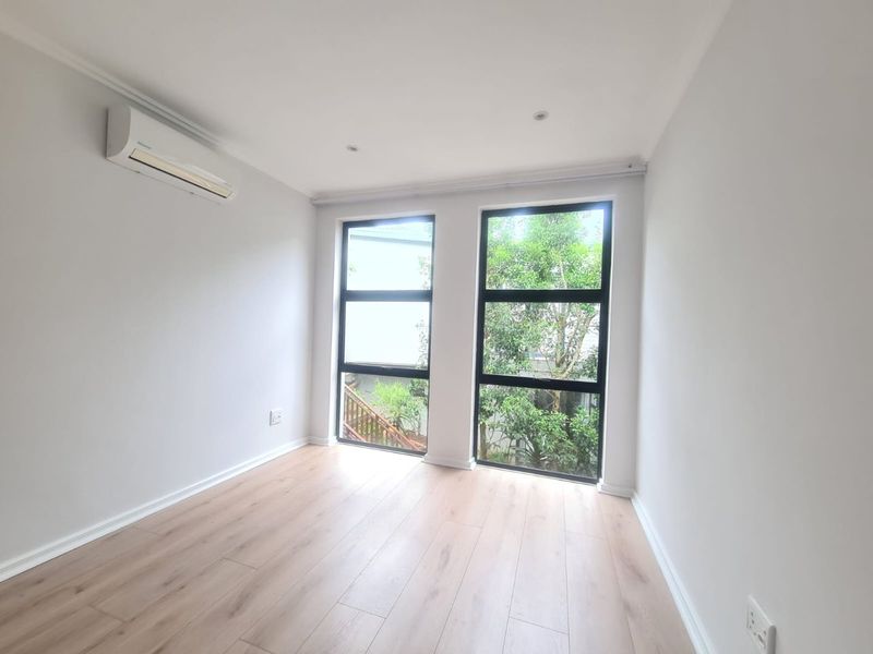 A bright, empty bedroom featuring modern laminate flooring, recessed downlights, and a wall-mounted split-system air conditioner with large windows overlooking greenery.