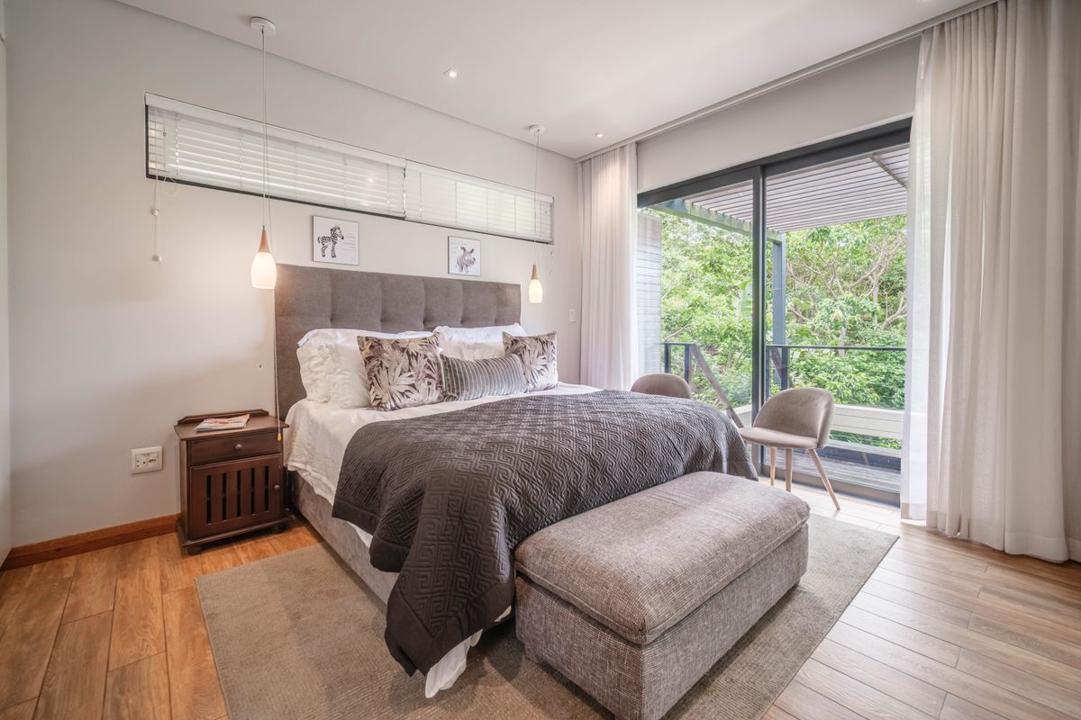 A well-appointed bedroom featuring timber-look tile flooring, modern pendant lighting, and large sliding glass doors opening to a private balcony with lush greenery views.