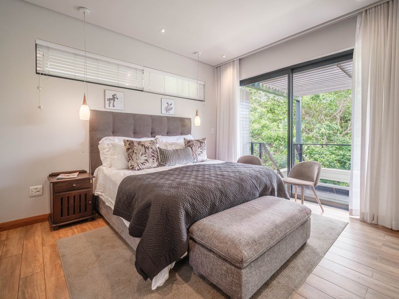 A well-appointed bedroom featuring timber-look tile flooring, modern pendant lighting, and large sliding glass doors opening to a private balcony with lush greenery views.