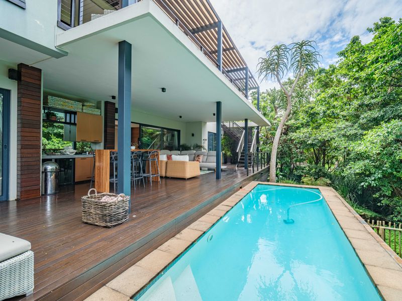 Spacious covered timber deck featuring an outdoor kitchen area, overlooking a swimming pool set within a lush, private garden environment.
