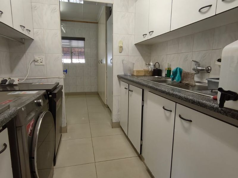 Functional laundry room featuring white built-in cabinetry, laminate countertops, a stainless steel sink, and tiled flooring with a connecting hallway.