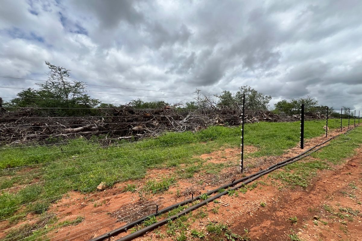 A view of the farm perimeter featuring an electric wire fence, irrigation piping, and cleared brush along the boundary line.