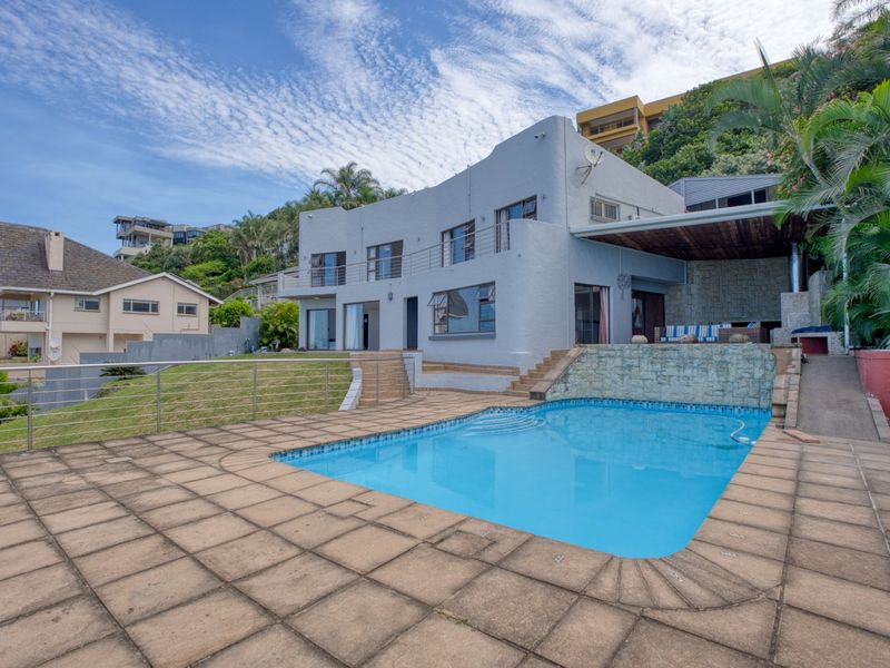 Rear exterior view featuring a swimming pool, stone water feature, paved deck, and a covered patio area set against a multi-level residential structure.
