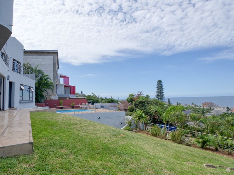 Rear exterior view featuring a landscaped lawn, tiled patio area, and an elevated swimming pool with ocean views in the distance.