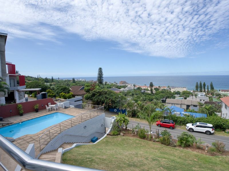 Elevated coastal view featuring a swimming pool, paved deck, and landscaped garden overlooking the ocean and surrounding residential area.