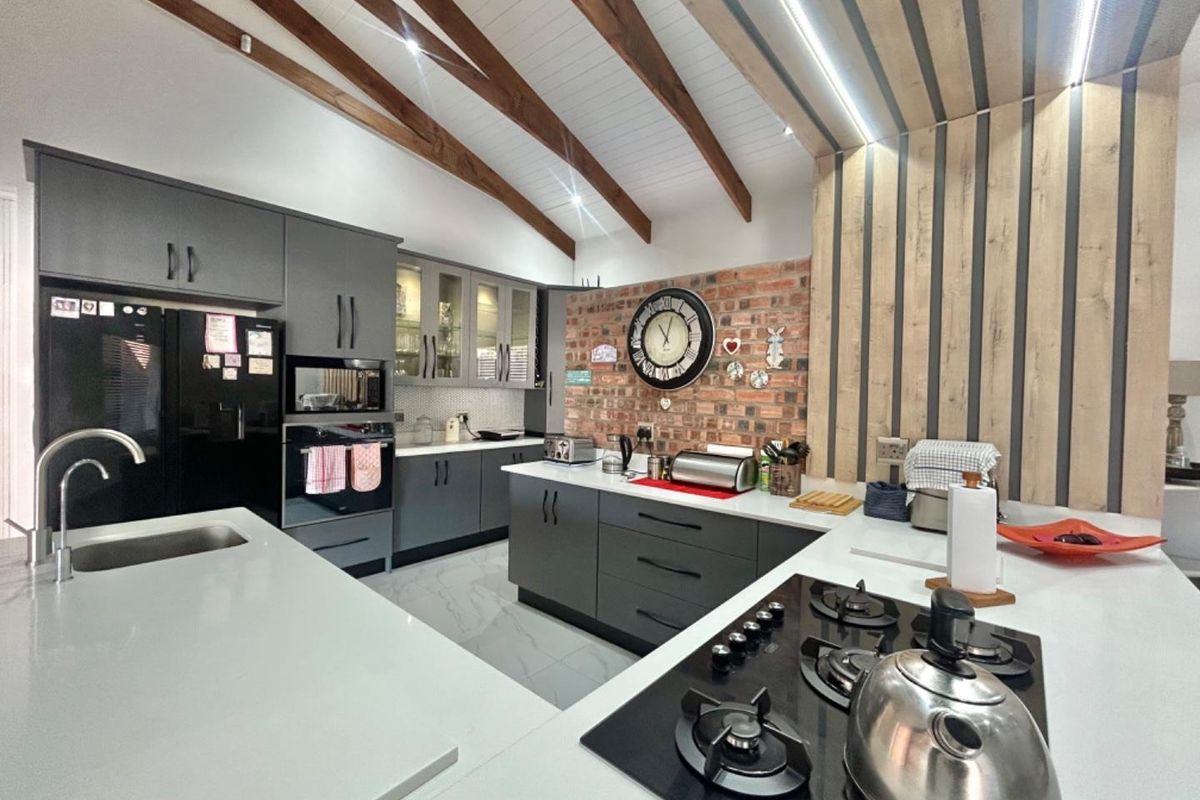 Modern kitchen featuring dark grey cabinetry, stone countertops, a gas hob, built-in oven, and a striking exposed brick feature wall under vaulted timber ceilings.