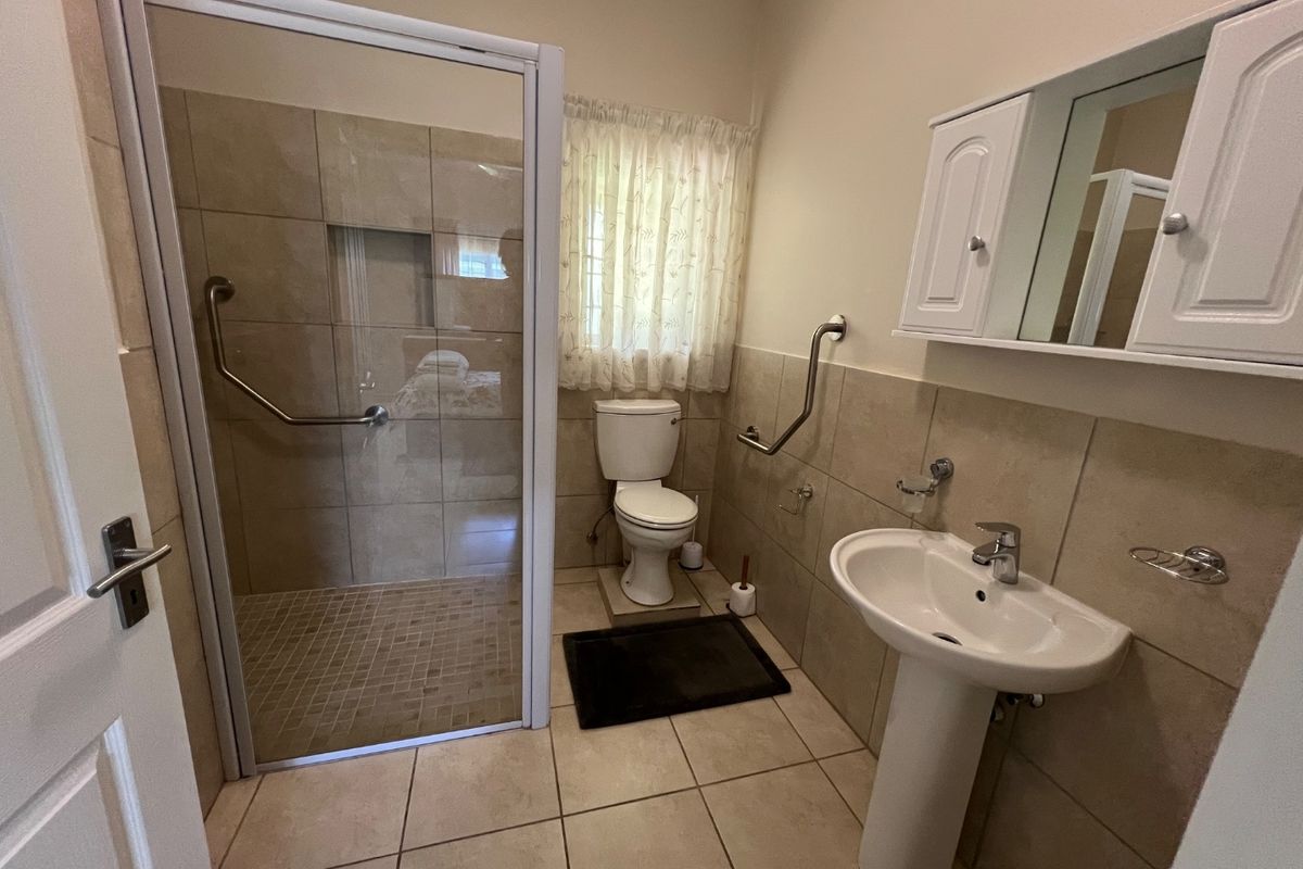 A functional bathroom featuring a walk-in shower with safety grab rails, a pedestal basin, and a wall-mounted medicine cabinet with neutral tiling.