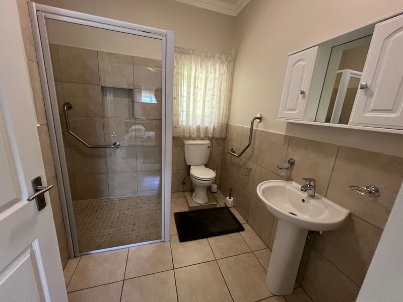 A functional bathroom featuring a walk-in shower with safety grab rails, a pedestal basin, and a wall-mounted medicine cabinet with neutral tiling.