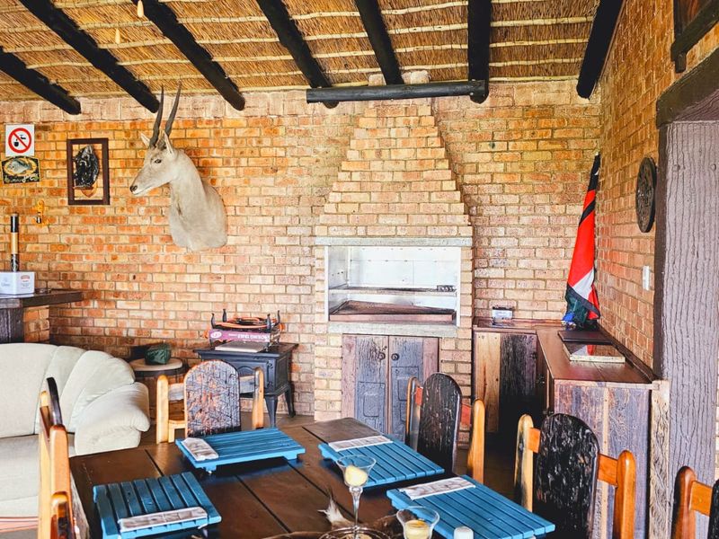 A rustic covered patio area featuring exposed brick walls, a traditional thatched roof with timber beams, and a built-in brick braai fireplace.