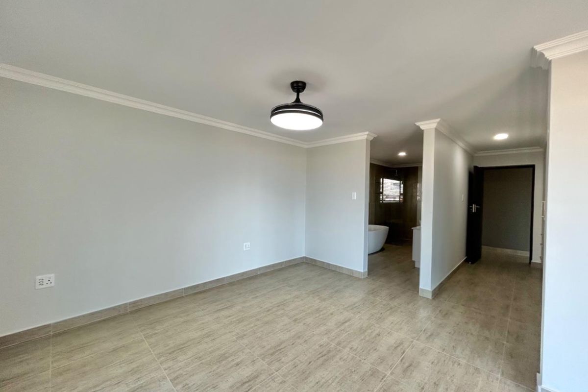 A spacious bedroom featuring neutral tiled flooring, crown molding, a modern ceiling fan, and an open-plan layout leading to an en-suite bathroom.