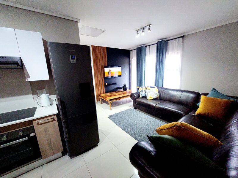 Modern open-plan living area featuring a compact kitchen with built-in appliances and a lounge space with feature wall paneling and tiled flooring.
