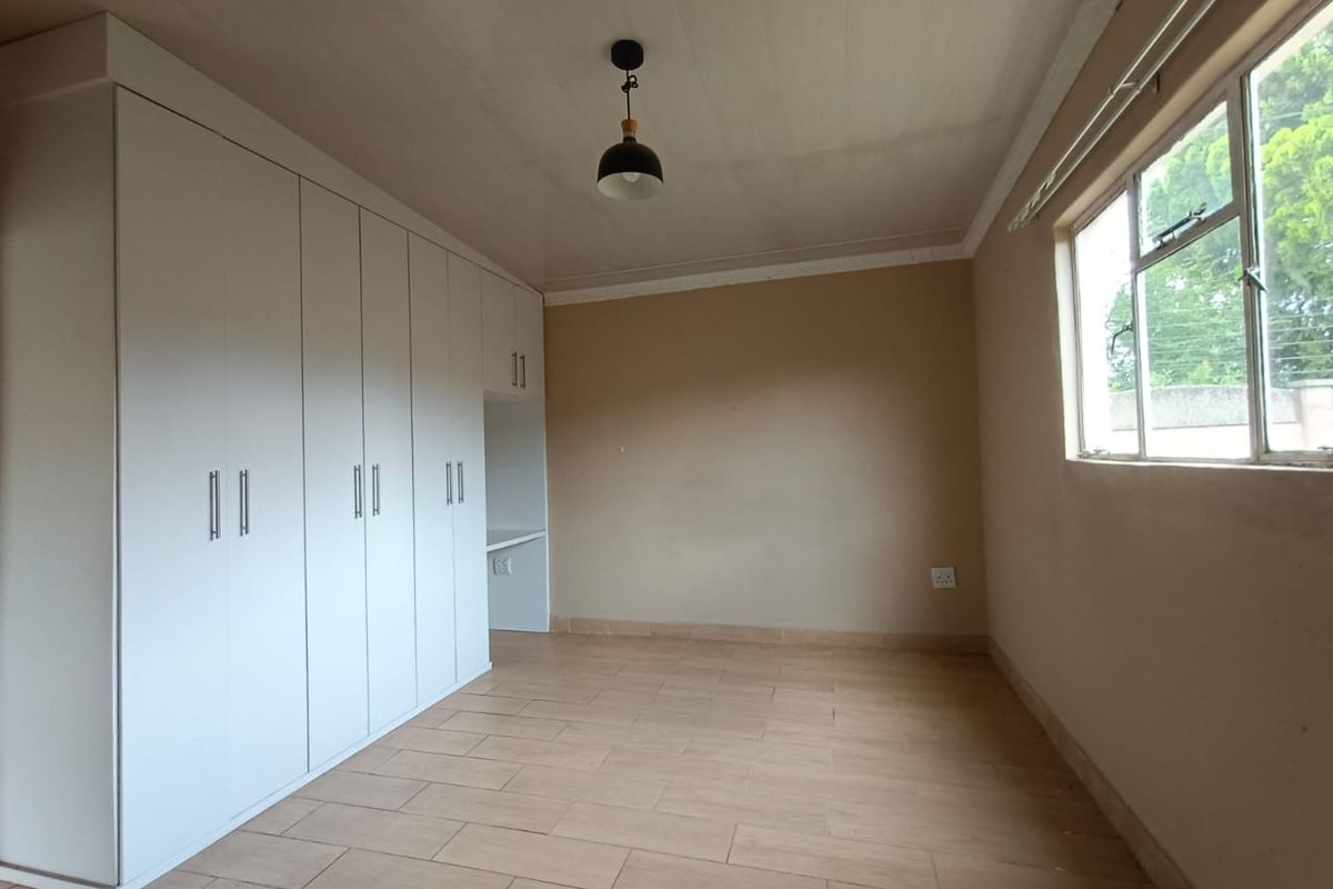 A bright, empty bedroom featuring neutral tiled flooring, built-in white cupboards with an integrated desk space, and a large window.