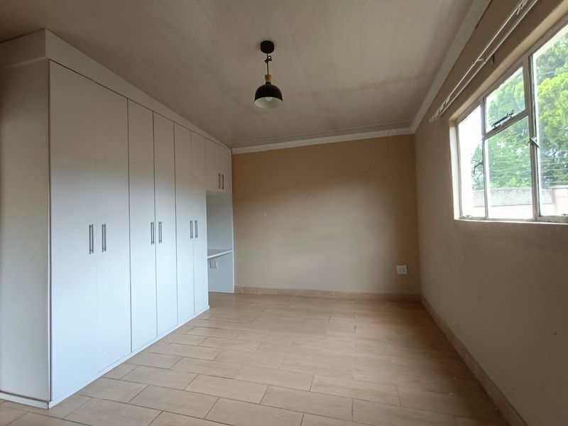 A bright, empty bedroom featuring neutral tiled flooring, built-in white cupboards with an integrated desk space, and a large window.
