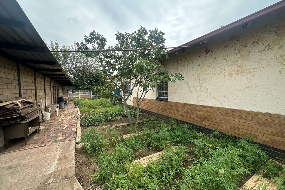 A rear garden area featuring a brick-paved walkway, vegetable patches, and a side building with a covered patio area.