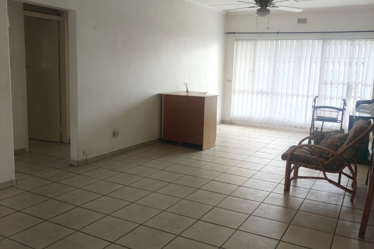 A spacious, open-plan living area featuring neutral floor tiles, a ceiling fan, and large windows with vertical blinds, offering a bright and functional layout.