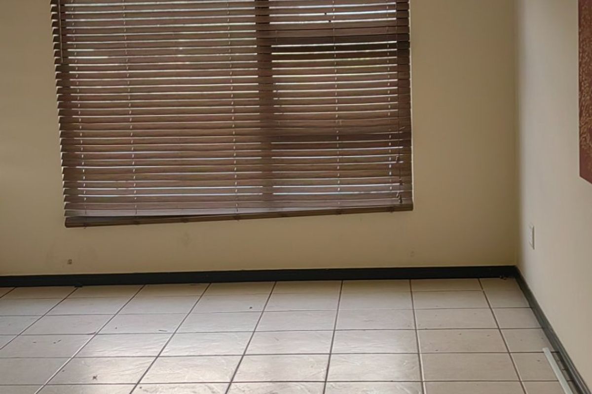 An empty bedroom featuring neutral tiled flooring, a large window with fitted venetian blinds, and a central ceiling light fixture.