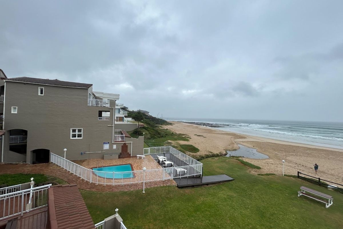 Elevated view of the apartment complex exterior, featuring a private swimming pool, paved patio area, and direct access to the beach and coastline.