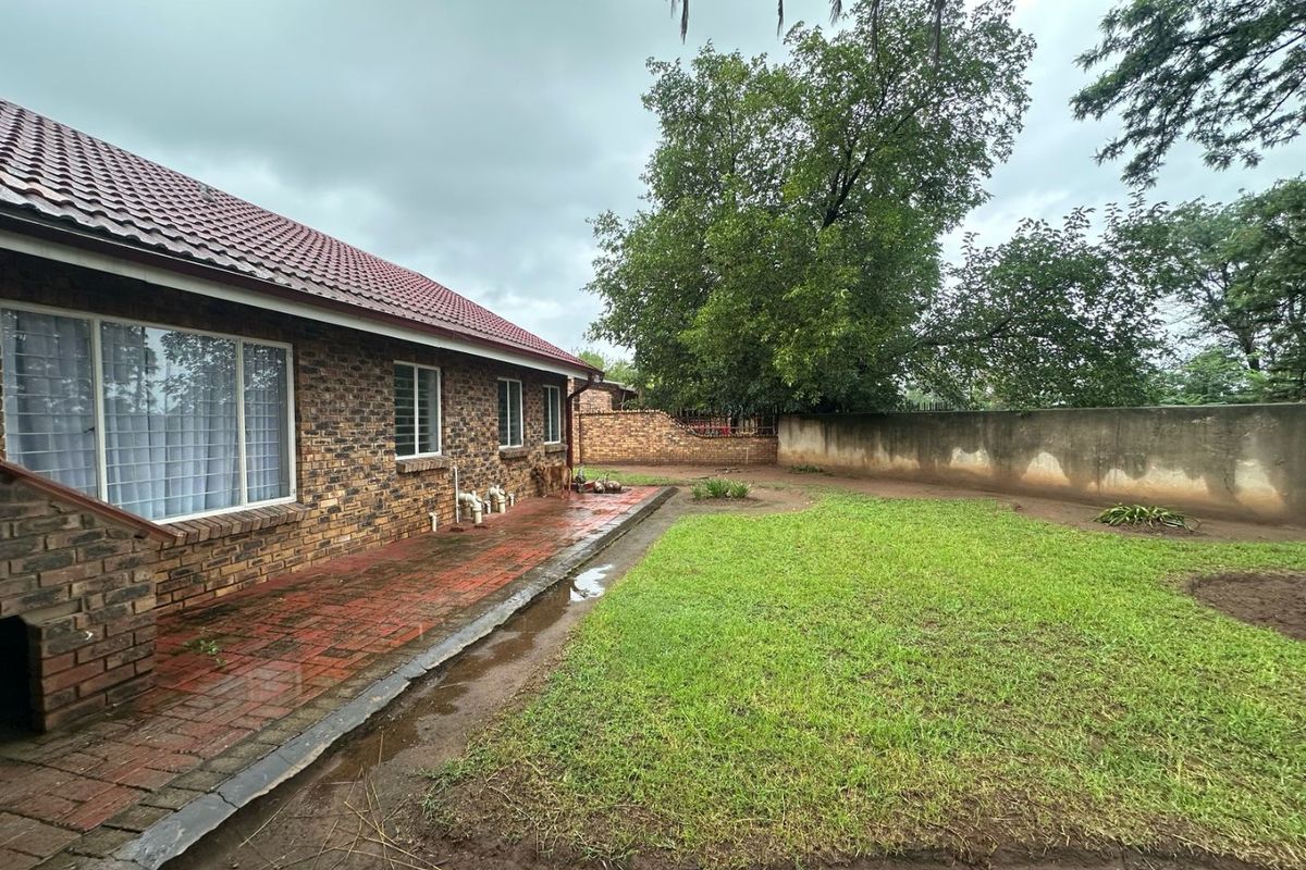 Rear exterior view of the house featuring a brick facade, tiled roof, paved patio area, and a spacious grass garden with a perimeter boundary wall.