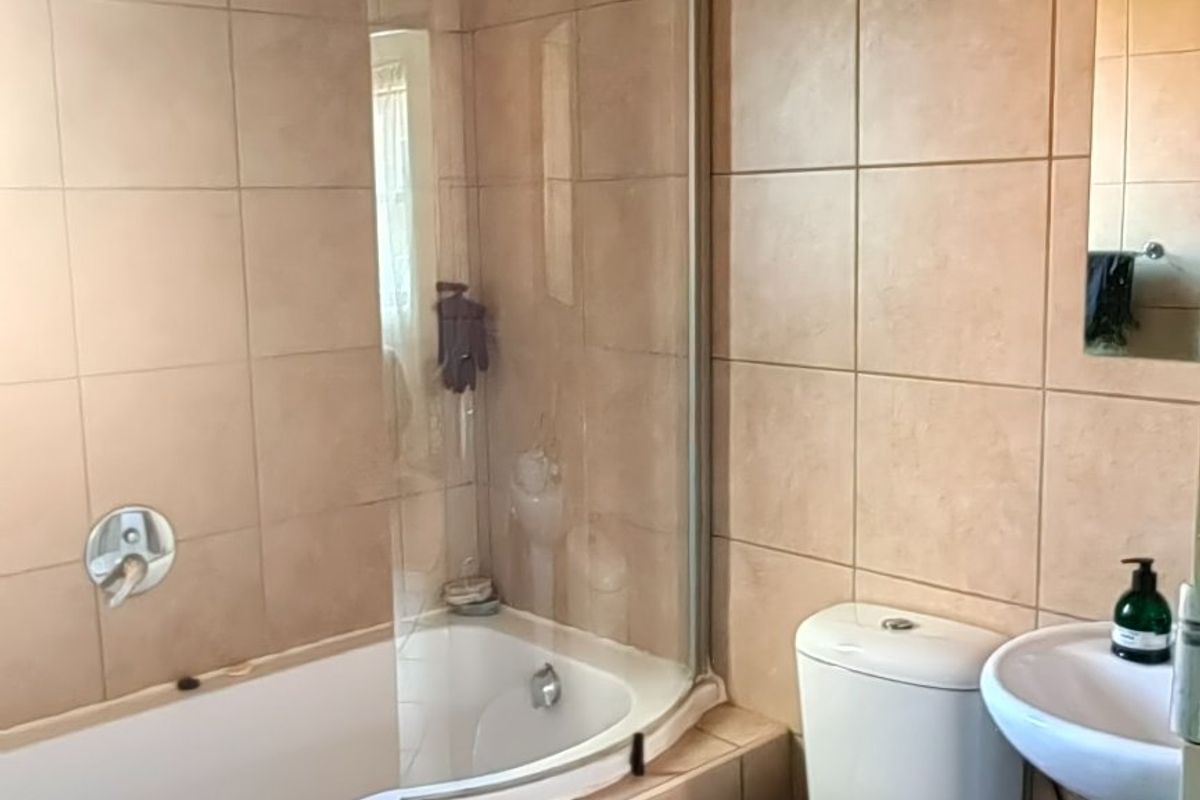 A functional bathroom featuring a corner bathtub with a glass screen, pedestal basin, and toilet, finished with neutral wall and floor tiles.