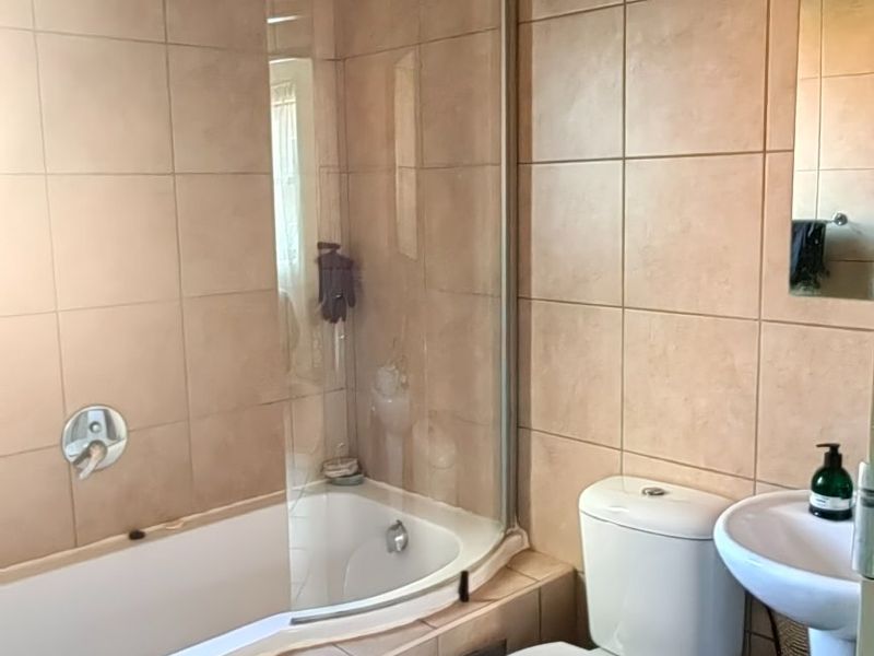 A functional bathroom featuring a corner bathtub with a glass screen, pedestal basin, and toilet, finished with neutral wall and floor tiles.