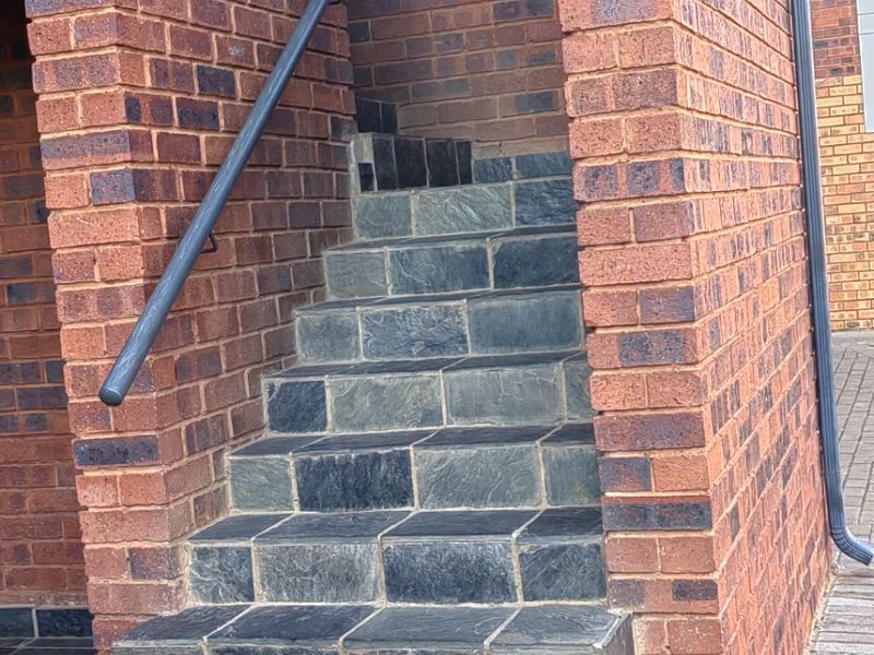 Exterior view of a brick building entrance featuring stone-tiled stairs and a metal handrail.