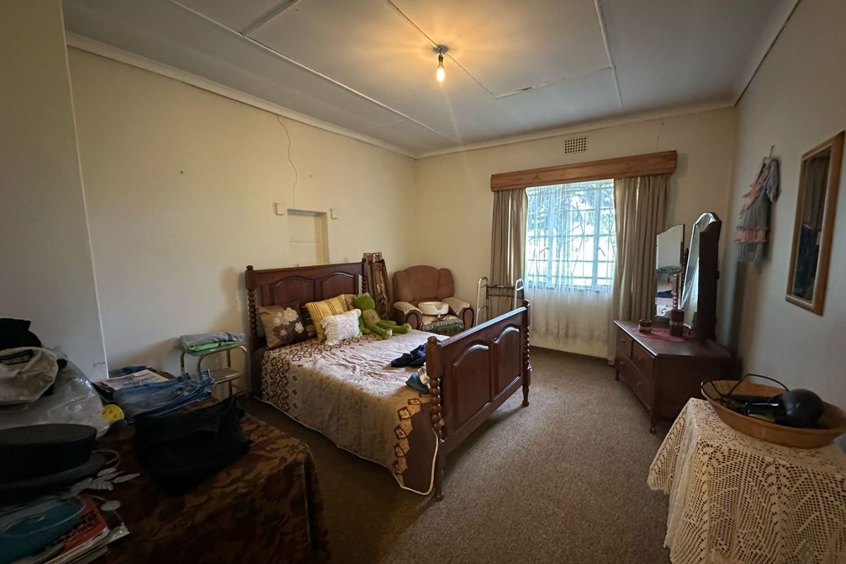 A spacious bedroom featuring carpeted flooring, a large window with wooden pelmet, and visible wall cracks requiring maintenance.