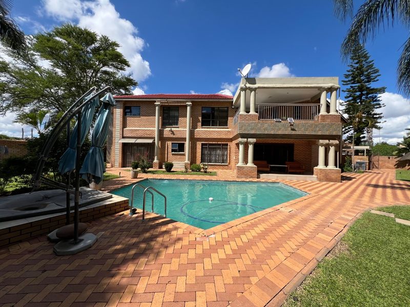 A spacious rear exterior view featuring a swimming pool, paved patio area, and a two-story brick house with a covered balcony and decorative columns.
