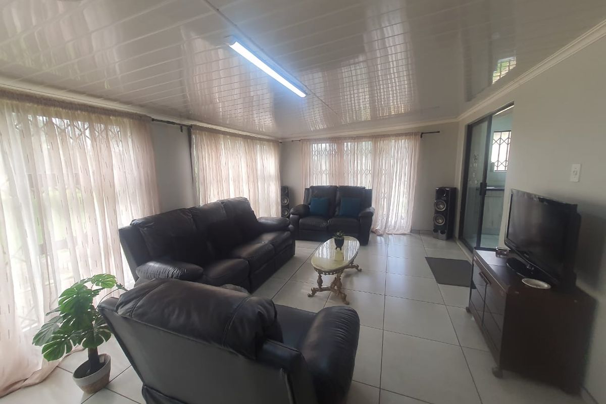 A spacious living room featuring large windows with security gates, tiled flooring, and a glossy panelled ceiling with integrated lighting.