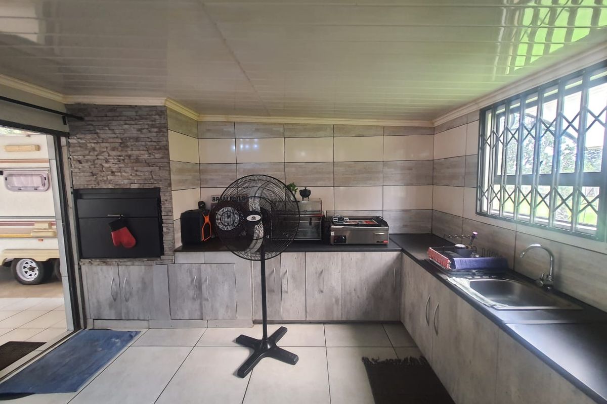 A functional outdoor kitchen area featuring built-in cabinetry, a stainless steel sink, and a built-in braai, finished with tiled surfaces and security bars.