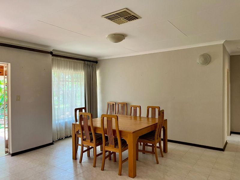 A spacious dining room featuring neutral tiled flooring, a ceiling vent, and natural light through a large window with curtain fittings.