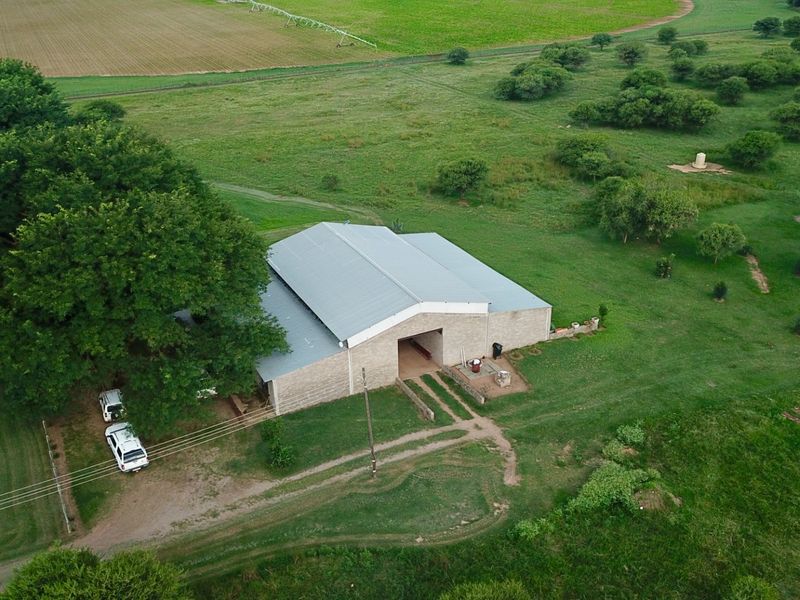 Aerial view of a large farm building with a metal roof, situated on expansive green pasture land with mature trees.