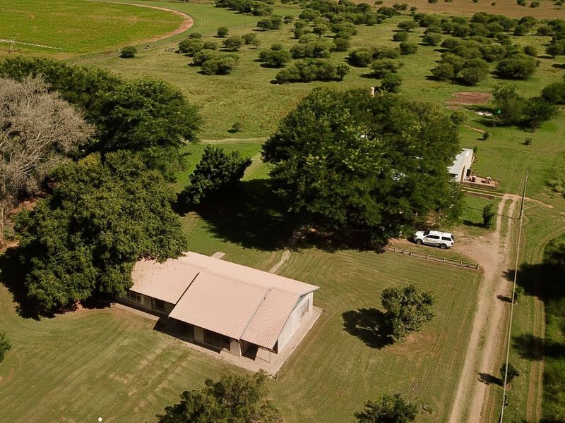 Aerial view of a farm property featuring a main dwelling with a metal roof, surrounded by expansive green fields and mature trees.