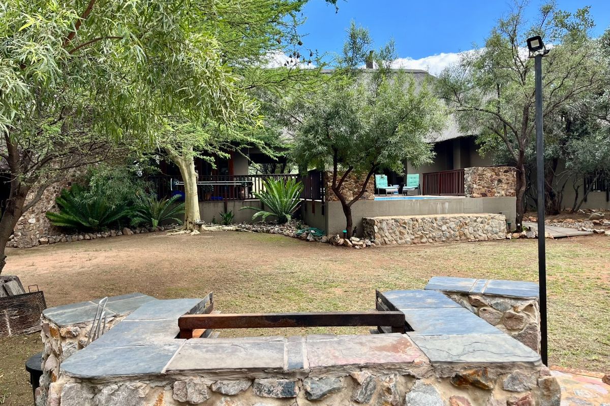 Rear garden view featuring a stone-built braai area, elevated patio with pool, and established trees in a natural bushveld setting.