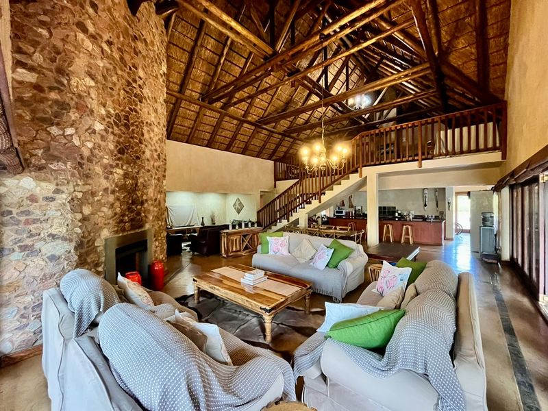 Spacious open-plan living area featuring a double-volume thatched roof, exposed timber trusses, a prominent stone fireplace, and a mezzanine level overlooking the space.