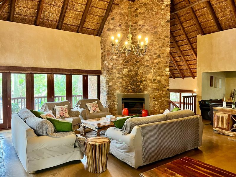 Spacious living room featuring a high vaulted thatched ceiling, a prominent floor-to-ceiling stone fireplace, and large windows overlooking the surrounding greenery.