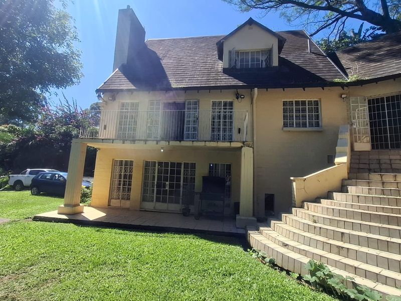 Rear view of a two-story house featuring a tiled patio, upper balcony, external staircase, and mature garden surroundings.