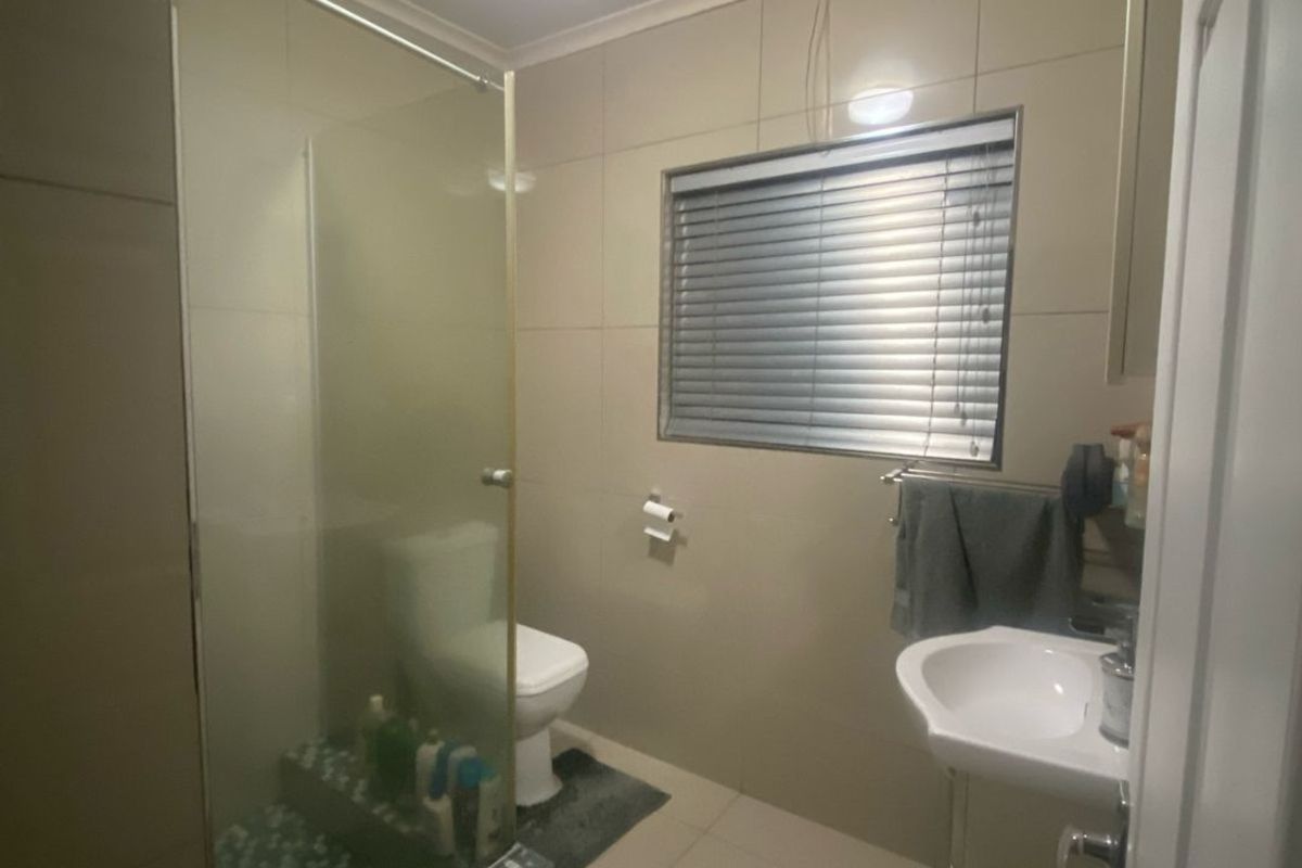 A functional bathroom featuring a glass-enclosed shower, pedestal basin, toilet, and neutral wall and floor tiling with a window for natural light.