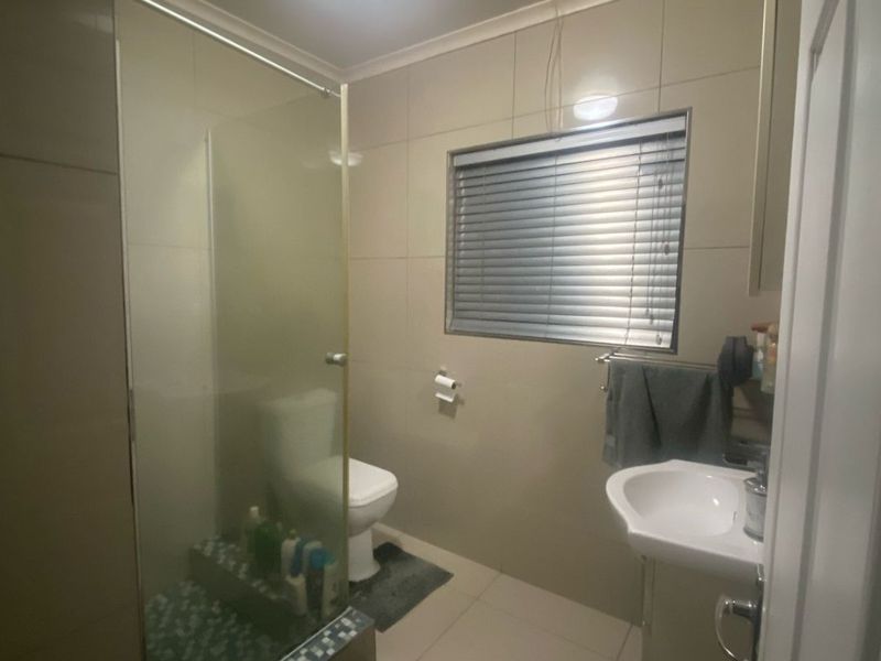 A functional bathroom featuring a glass-enclosed shower, pedestal basin, toilet, and neutral wall and floor tiling with a window for natural light.