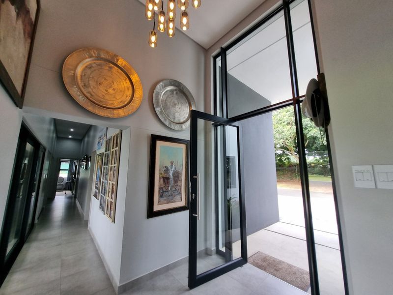 Spacious modern entrance hall featuring high ceilings, large glass pivot door, and contemporary pendant lighting, leading into a tiled hallway.