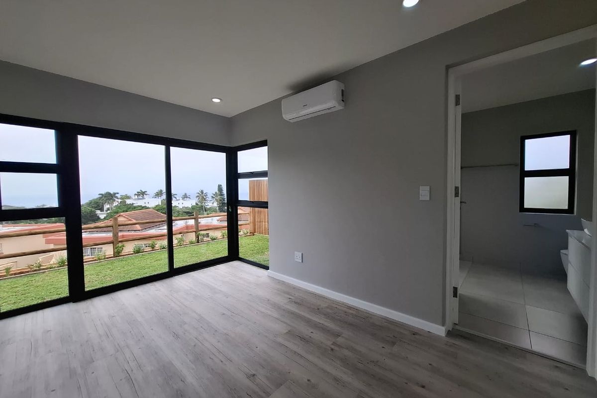 Modern bedroom featuring light wood-look flooring, large black-framed windows with garden views, a wall-mounted air conditioning unit, and an en-suite bathroom.