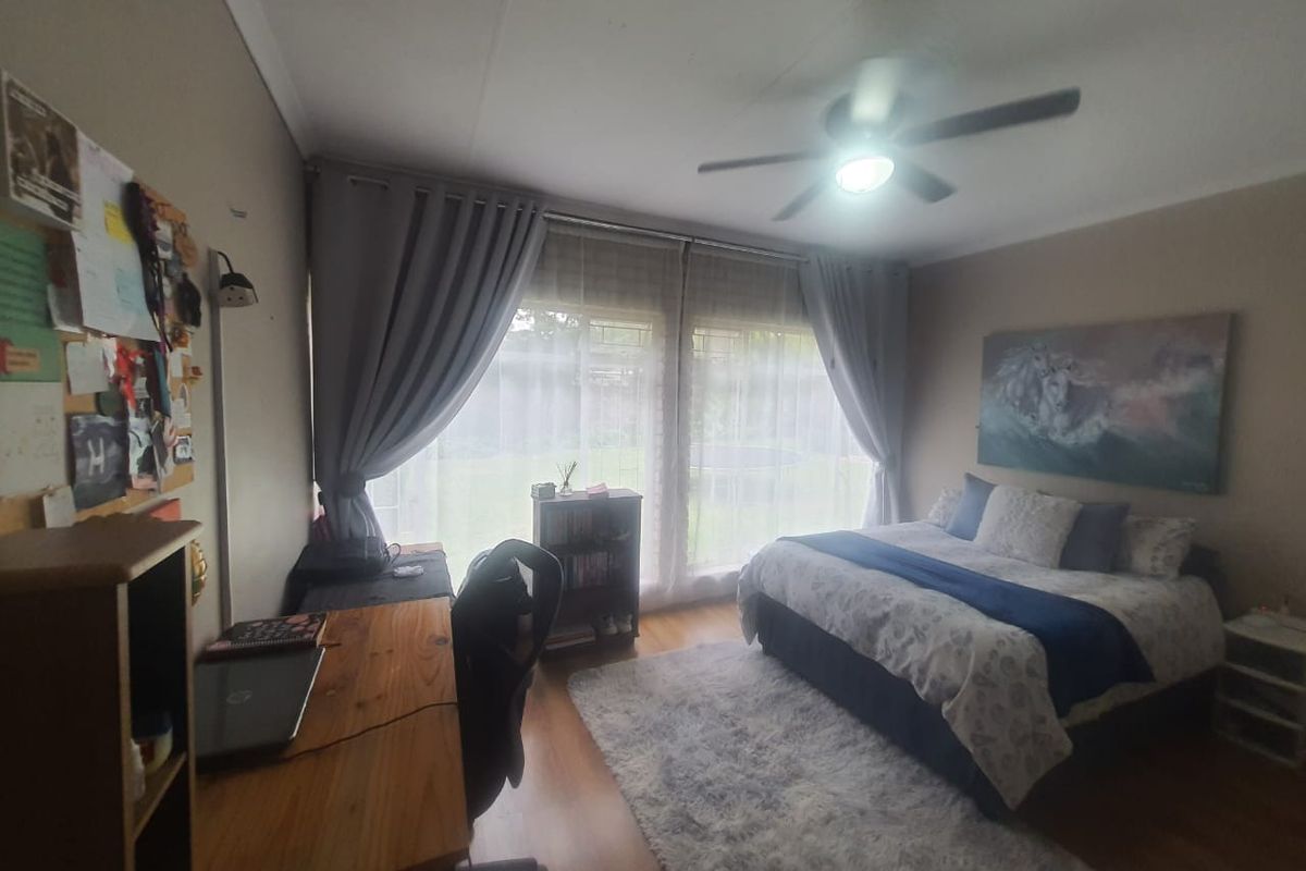 A well-lit bedroom featuring laminate flooring, a ceiling fan, and large windows with curtains overlooking the garden.
