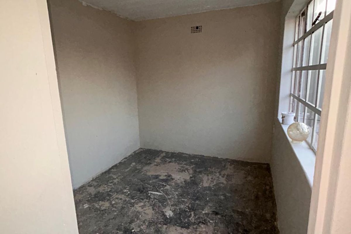 An empty, unfinished bedroom space requiring significant renovation, featuring a steel-framed window and exposed floor surface.