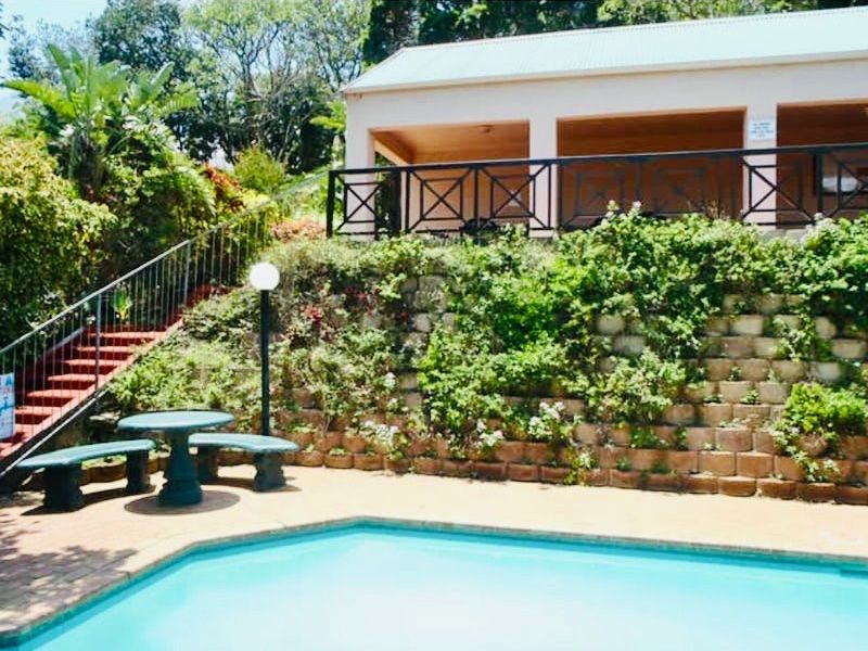 Outdoor pool area featuring a paved patio, retaining wall with landscaping, and a metal balustrade leading to an elevated covered terrace.