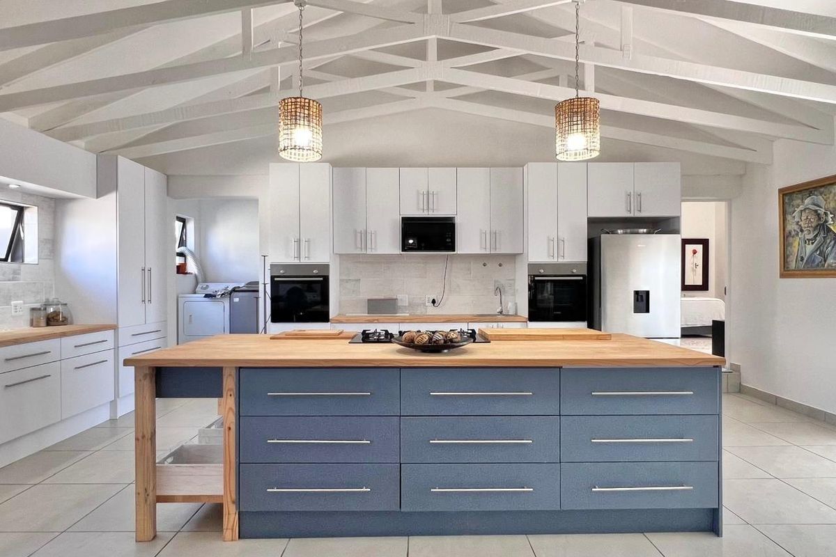 Modern open-plan kitchen featuring a large central island with a wooden countertop, built-in ovens, white cabinetry, and exposed vaulted ceiling beams.