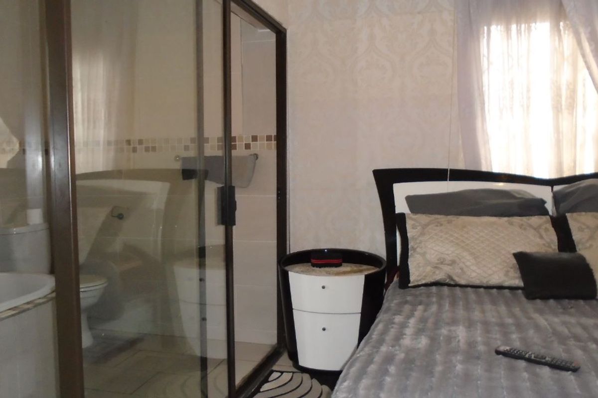 A bedroom featuring an en-suite bathroom with a glass-enclosed shower and toilet, finished with neutral wall coverings and tiled flooring.