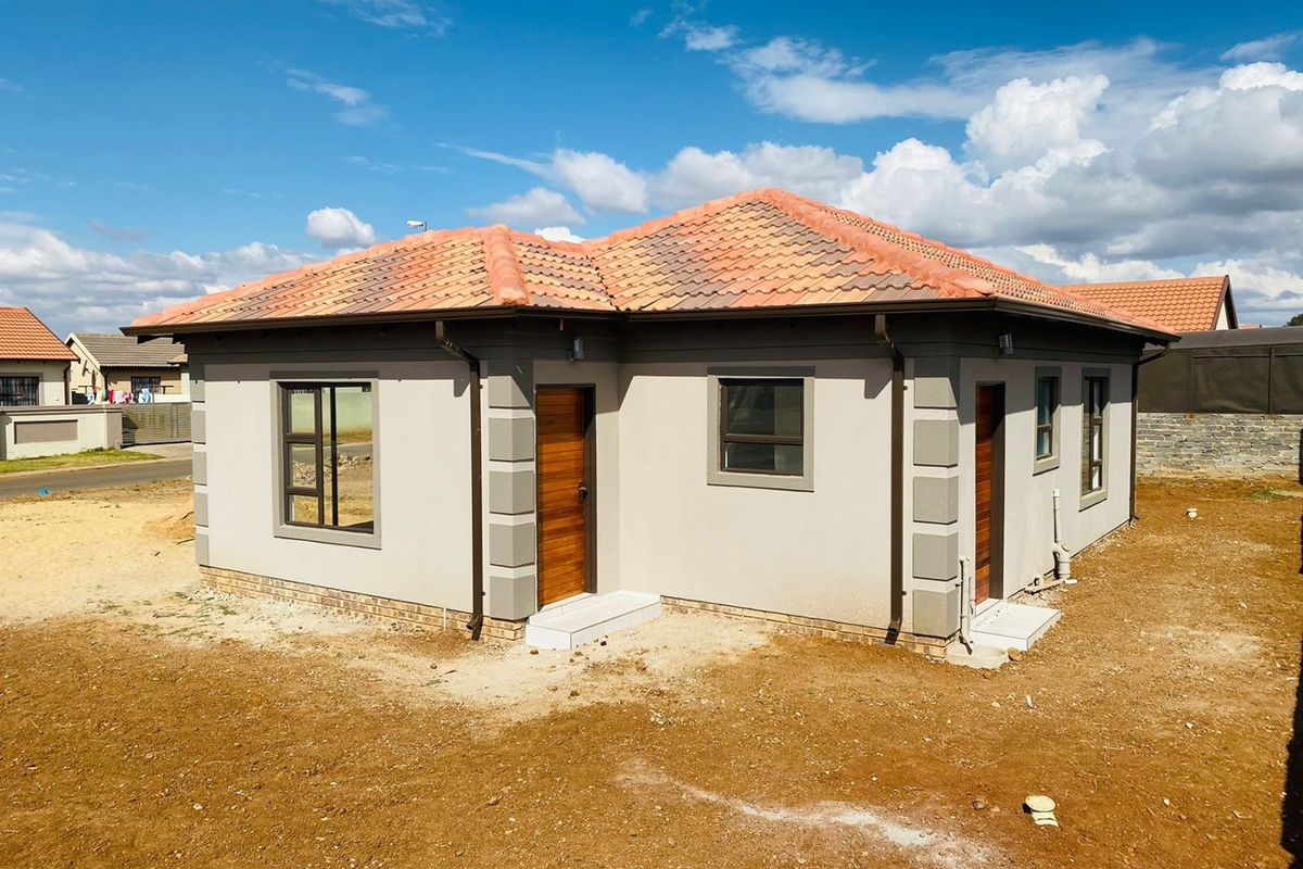 A modern single-story house featuring a tiled hip roof, neutral exterior plaster, and wooden entrance doors, set on an undeveloped plot.